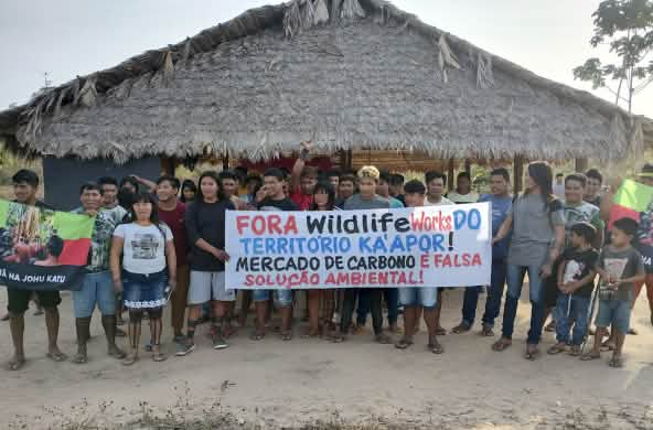 Eine Gruppe von etwa 30 Indigenen protestiert mit einem Banner vor einer mit Palmenwedeln überdachten Hütte. Aufschrift des Banners: "Wildlife Works raus aus dem Territorium der Ka'apor. Der Kohlenstoffhandel ist eine falsche Umweltlösung"